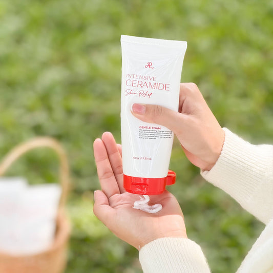A person's hands holding a white tube of Intensive Ceramide Skin Relief Gentle Foam sunscreen, with white cream being squeezed onto their palm, set against a soft-focus green outdoor background.