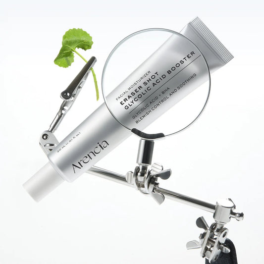 A silver tube of Arencia Eraser Shot Glycolic Acid Booster facial moisturizer is mounted on a metal stand with a magnifying glass and a small green leaf prop against a clean white background.
