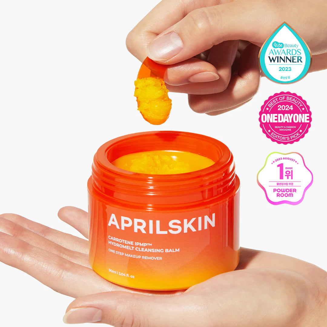 A person's hands holding an open jar of Aprilskin Carrotene IPMP Hydramelt Cleansing Balm, with a focus on the bright orange product and the brand's logo.