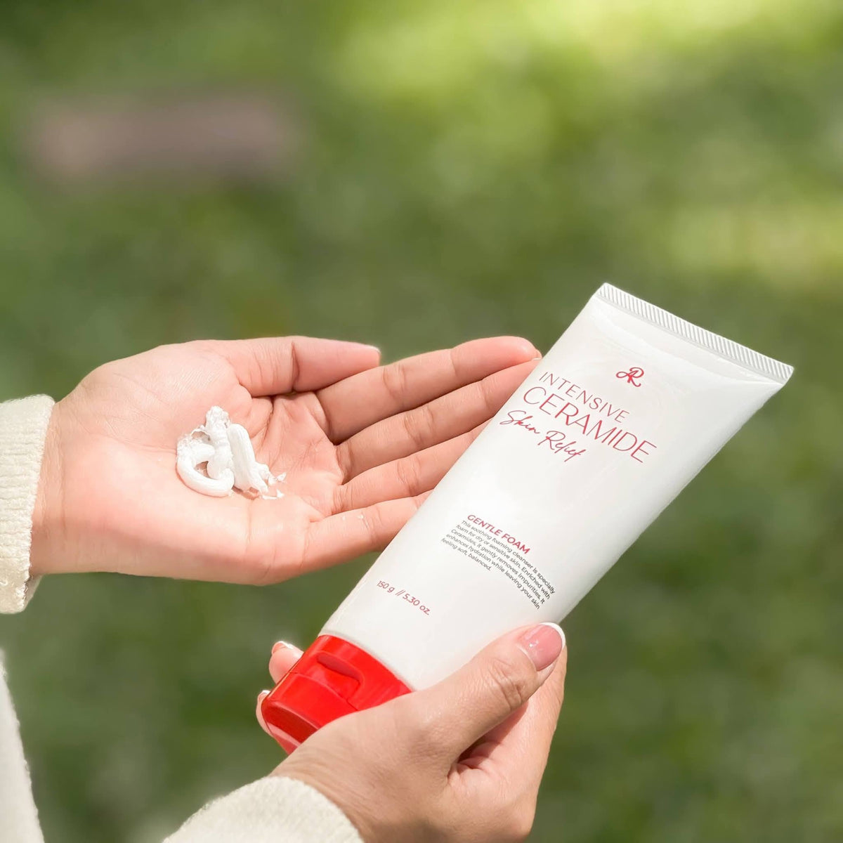 A person holds a white tube of Ceramide Ato Skin Relief Intensive Ceramide Cream with a red cap, with a dollop of the white cream in their other hand, set against a soft, blurred green outdoor background.