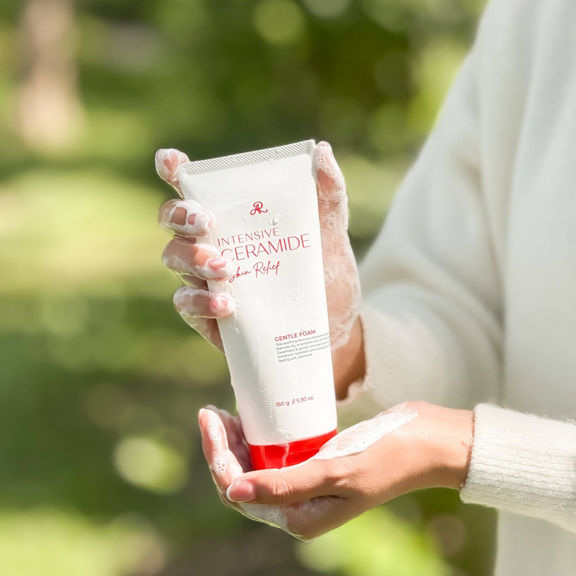 A person in a white sweater holds a white tube of John LaRue Intensive Ceramide Skin Relief Gentle Flush moisturizer with a red cap, covered in white cream, against a blurred green outdoor background.