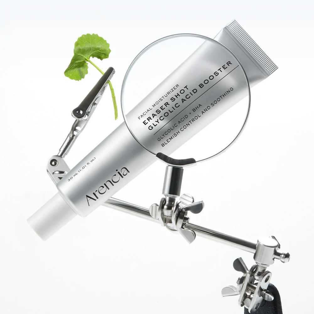 A silver tube of Arencia Eraser Shot Glycolic Acid Booster facial moisturizer is mounted on a metal stand with a magnifying glass and a small green leaf prop against a clean white background.