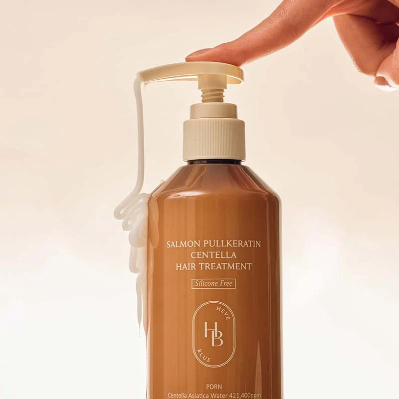 A bottle of hair treatment with a hand pressing the pump, showing a white cream dripping out. The bottle is a warm salmon-brown color with white text and logo, set against a clean, light beige background.