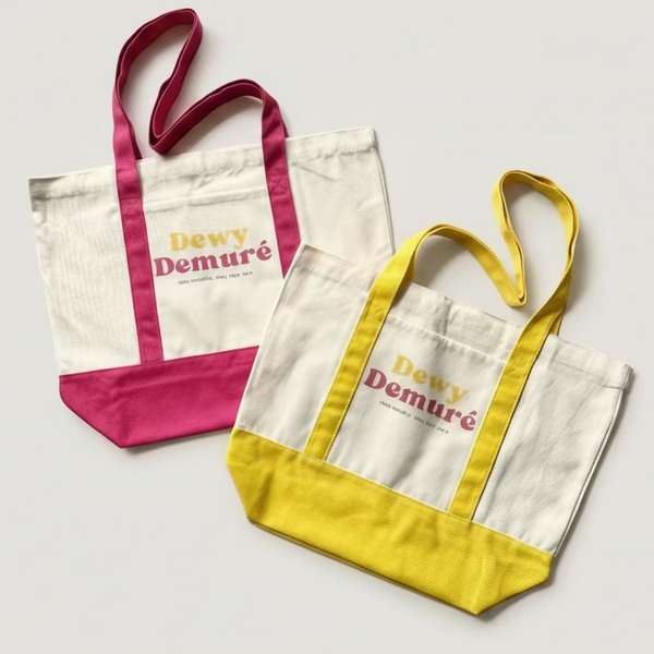 Two tote bags with 'Dewy Demure' branding on a tiled floor.