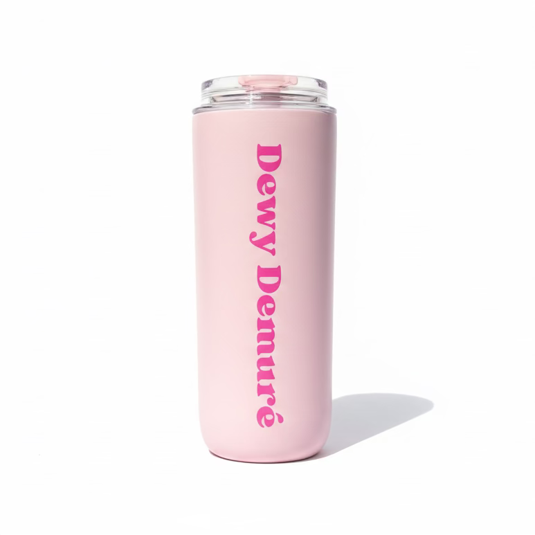 Pink tumbler with 'Dewy Demure' text held in a hand outdoors.