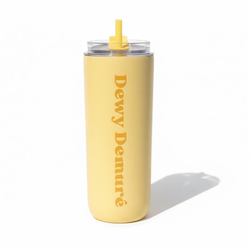 Hand holding a beige tumbler with 'Dewy Demure' text against a grassy outdoor background