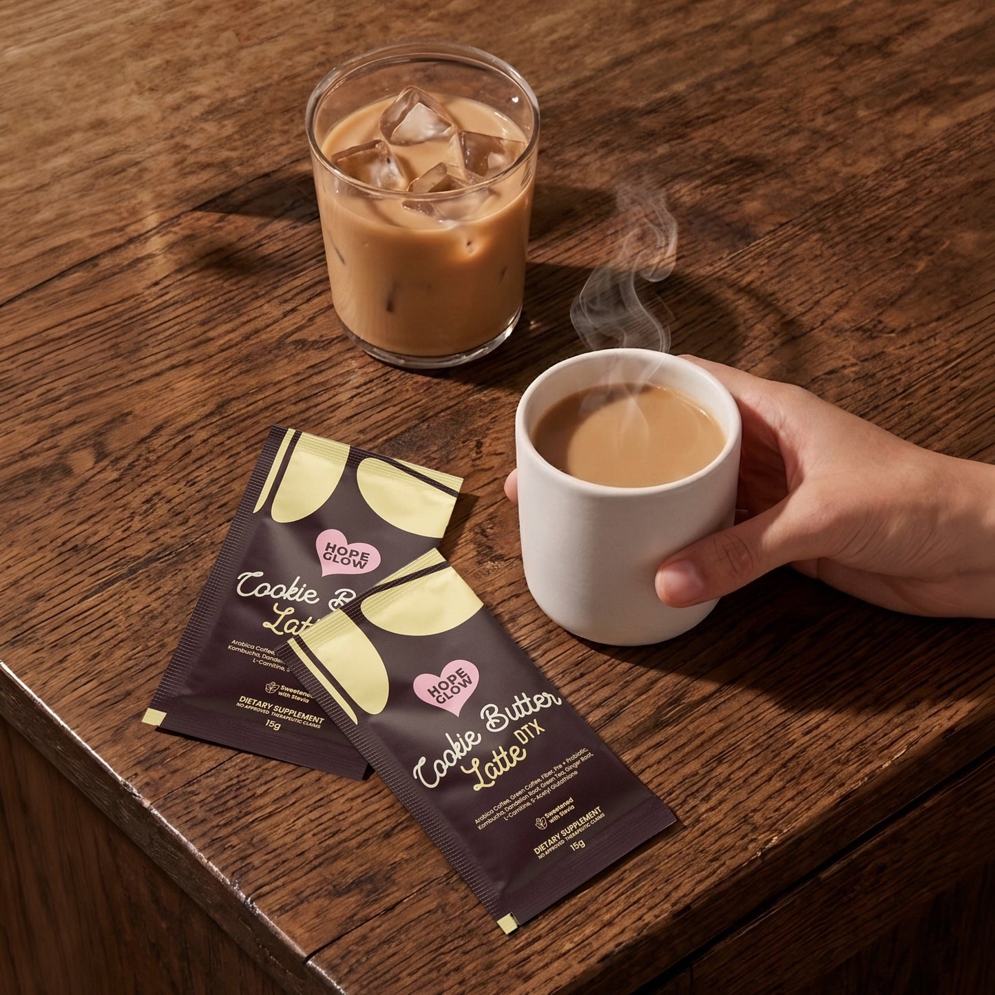 Hand holding a steaming cup of coffee next to an iced coffee and two packets on a wooden surface